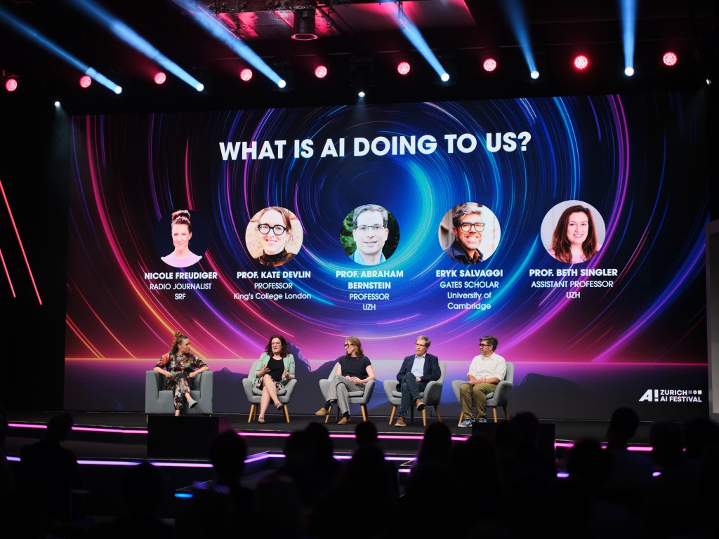 This image shows a stage with a large screen behind it displaying the text "WHAT IS AI DOING TO US?" and images of five individuals with their names and titles. The stage is illuminated with colorful lights and has a modern design. Five people are seated on chairs on the stage, likely participating in a discussion or panel. The audience is seated…